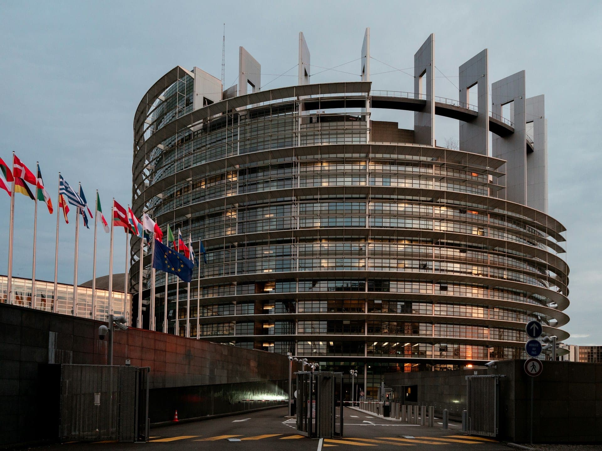 Clădirea Parlamentului European de la Strasbourg în amurg, cu drapelele UE și ale statelor membre pe catarge în față.